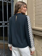 A woman wearing a dark navy blue blouse with white crochet lace sleeve panels and cuffs, shown from the back in an outdoor setting next to a beige brick wall.