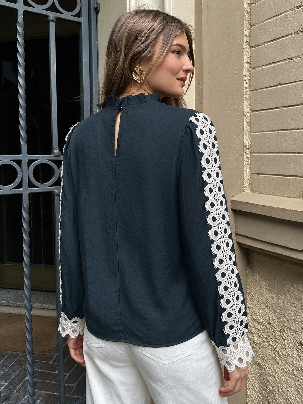 A woman wearing a dark navy blue blouse with white crochet lace sleeve panels and cuffs, shown from the back in an outdoor setting next to a beige brick wall.