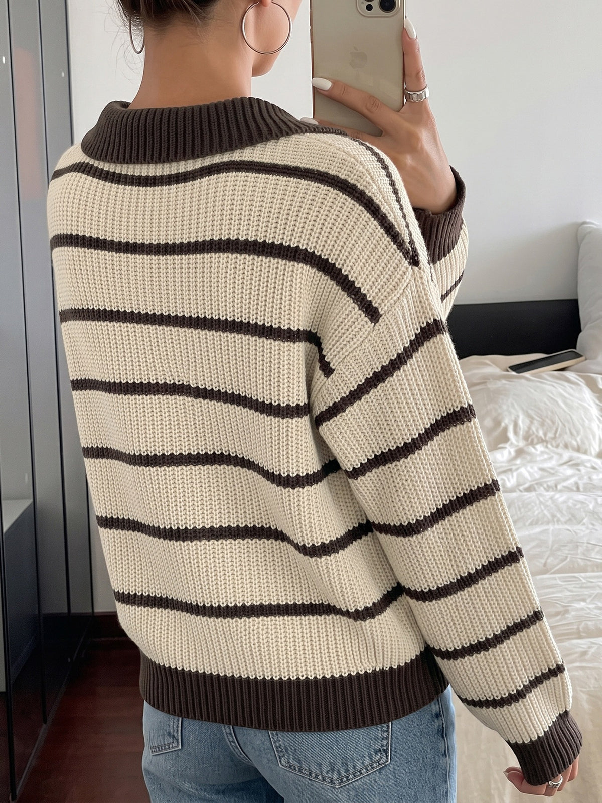 A person taking a mirror selfie in a bedroom, wearing a cream and dark brown horizontally striped knit sweater with a crew neck, dropped shoulders, and ribbed cuffs and hem.