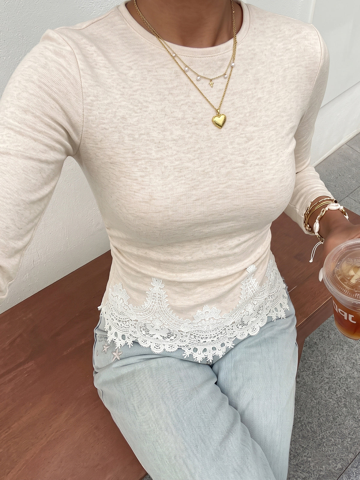 A woman wearing a fitted long-sleeve cream top with white lace trim at the hem, paired with light blue jeans and layered gold necklaces, seated on a wooden bench.