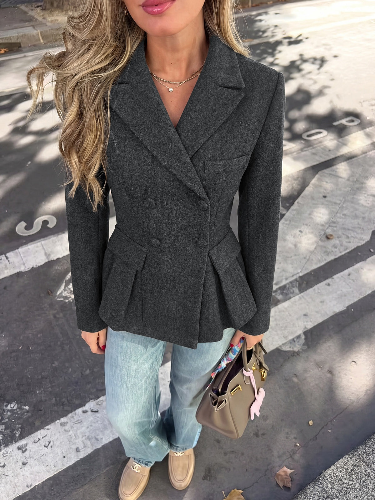 A woman wearing a dark gray double-breasted blazer with a peplum waist, paired with light blue jeans and beige loafers, holding a brown handbag on a city street.