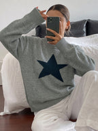 A person sitting on a bed taking a mirror selfie, wearing a heather gray knit sweater with a large dark navy blue star on the front, paired with white pants.