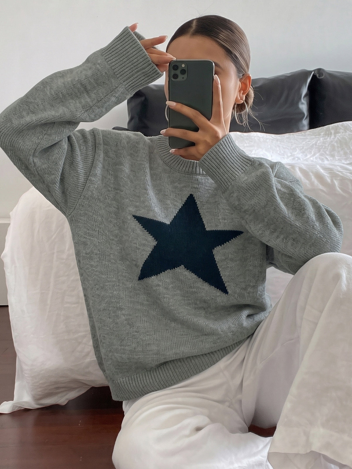 A person sitting on a bed taking a mirror selfie, wearing a heather gray knit sweater with a large dark navy blue star on the front, paired with white pants.