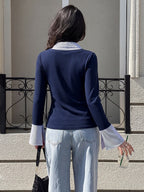 A woman viewed from behind wearing a navy long-sleeve top with contrasting white collar and cuffs, paired with light-wash jeans, standing outdoors in front of a building wall and black metal railing.