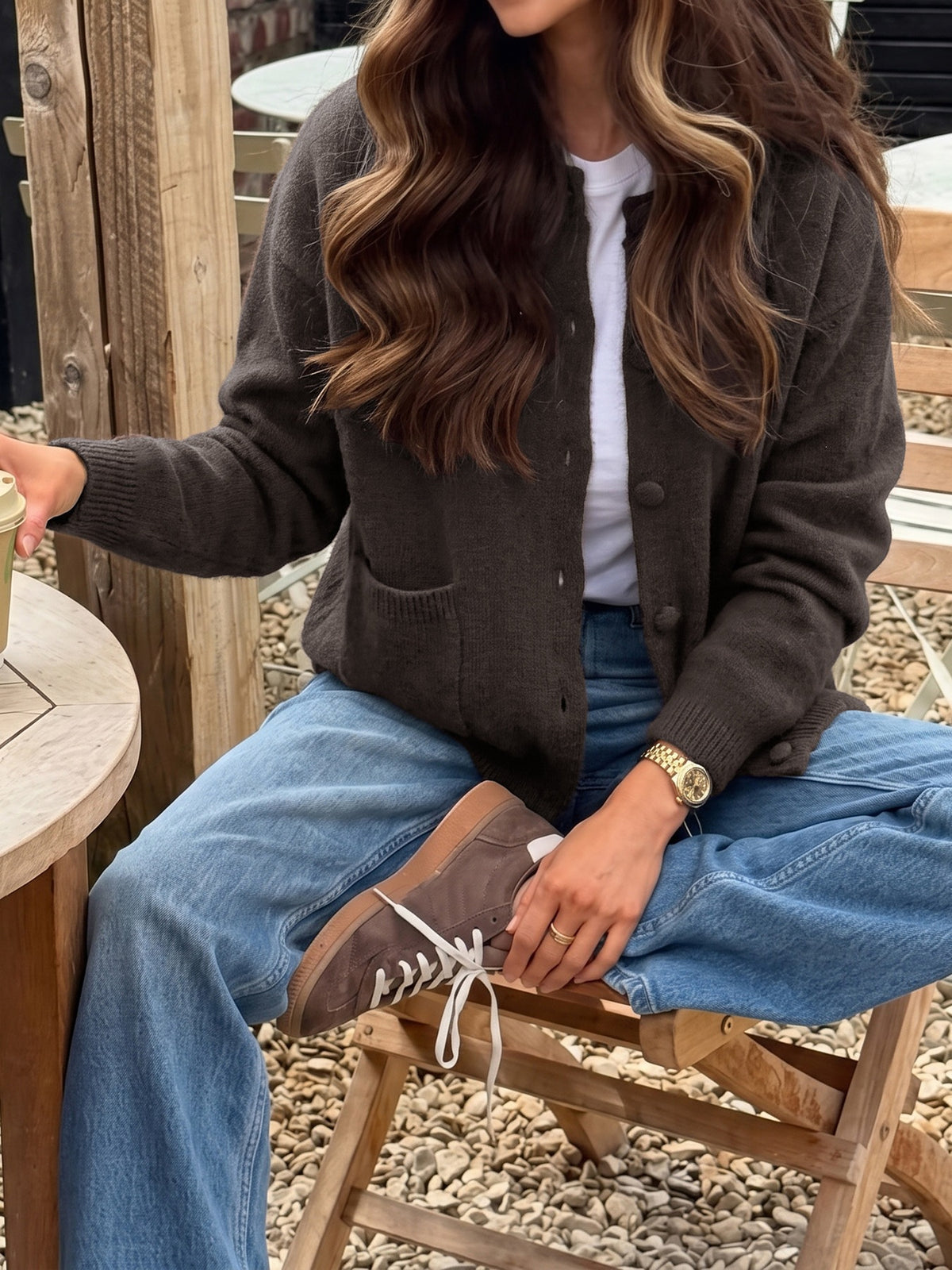 A woman seated outdoors on a wooden stool wearing a dark brown button-front cardigan over a white top, blue jeans, and brown lace-up shoes.