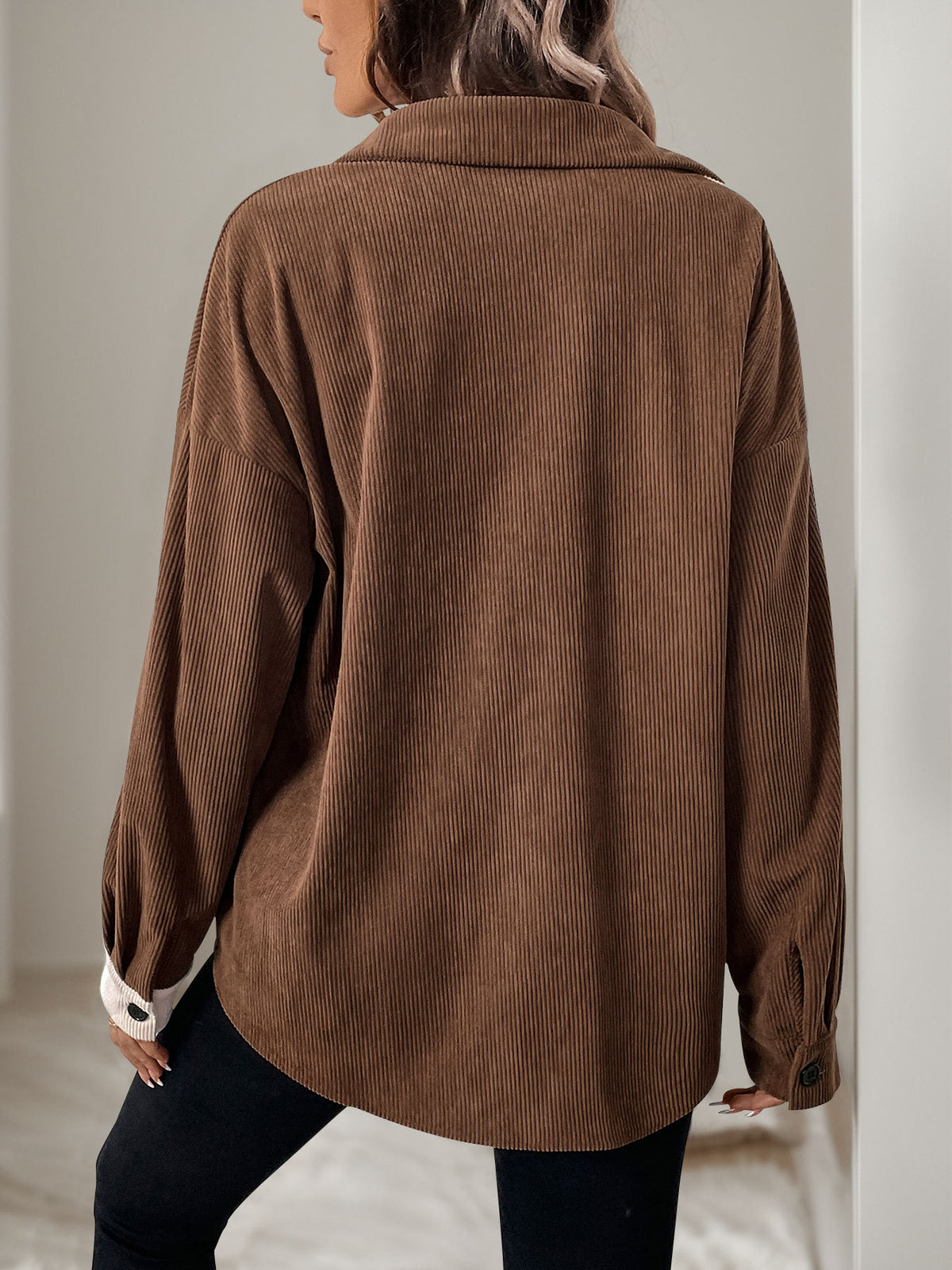 A woman shown from the back wearing an oversized brown corduroy button-up shirt with dark pants, standing in a neutral indoor setting.