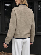A woman shown from the back wearing a beige knit mock-neck sweater with dark navy ribbed trim at the collar, cuffs, and hem, paired with white wide-leg pants, set against a dark building facade.