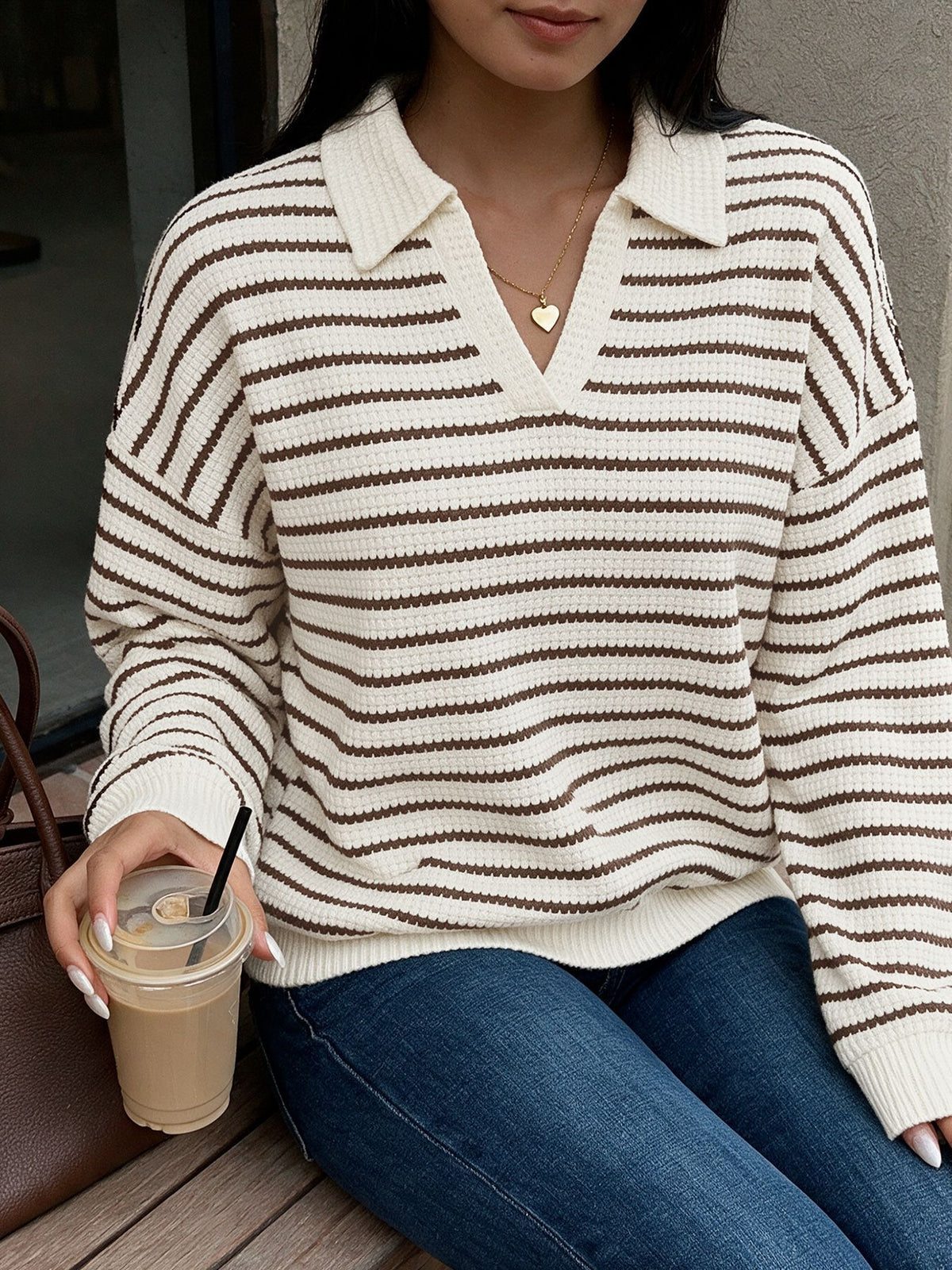 A woman wearing a cream and brown horizontally striped knit polo sweater with a V-neck, paired with blue jeans, holding an iced coffee.