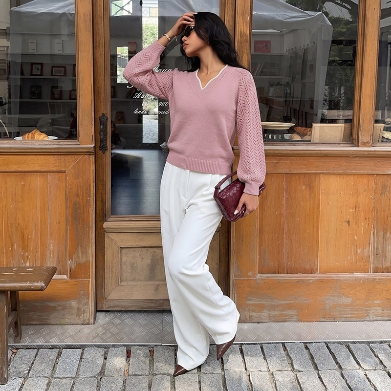 A woman stands in front of a rustic wooden storefront wearing a dusty pink knit sweater with white trim, white wide-leg trousers, and brown pointed shoes, while holding a burgundy handbag.