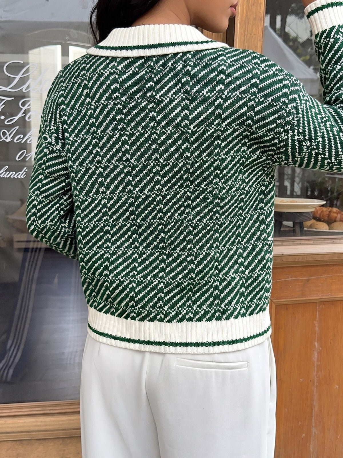 A person shown from the back wearing a green and white patterned knit sweater with ribbed collar, hem, and cuffs, paired with white pants in an indoor setting.