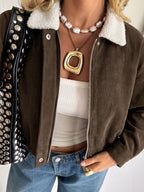 A close-up of a person wearing a brown corduroy jacket with a white sherpa collar, layered over a white ribbed tube top and blue jeans. The person is accessorized with layered necklaces and a black studded shoulder bag.