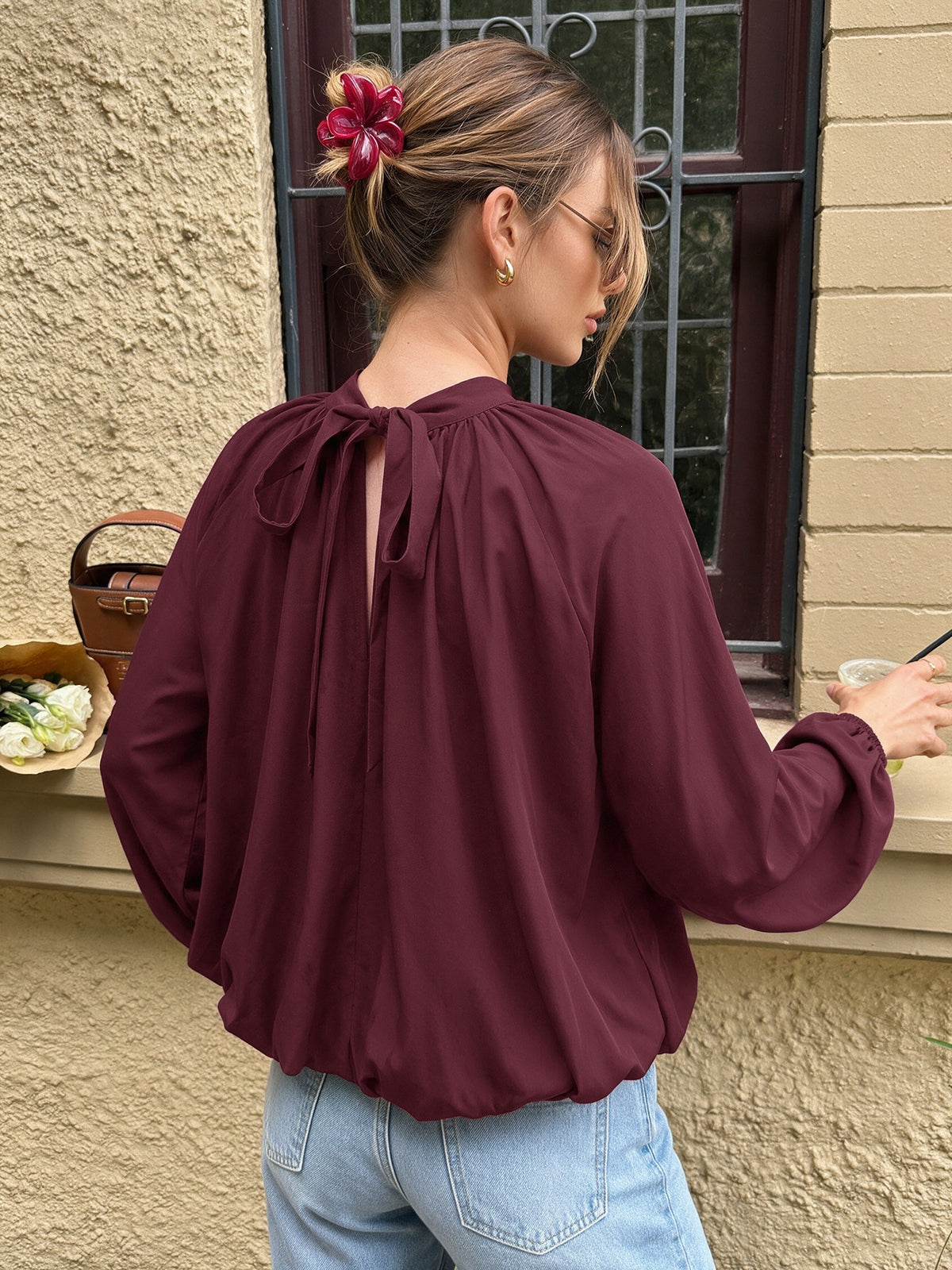 A woman wearing a long-sleeve burgundy blouse with a tie-back neckline, paired with light blue jeans, standing outdoors in front of a beige stucco wall and window.