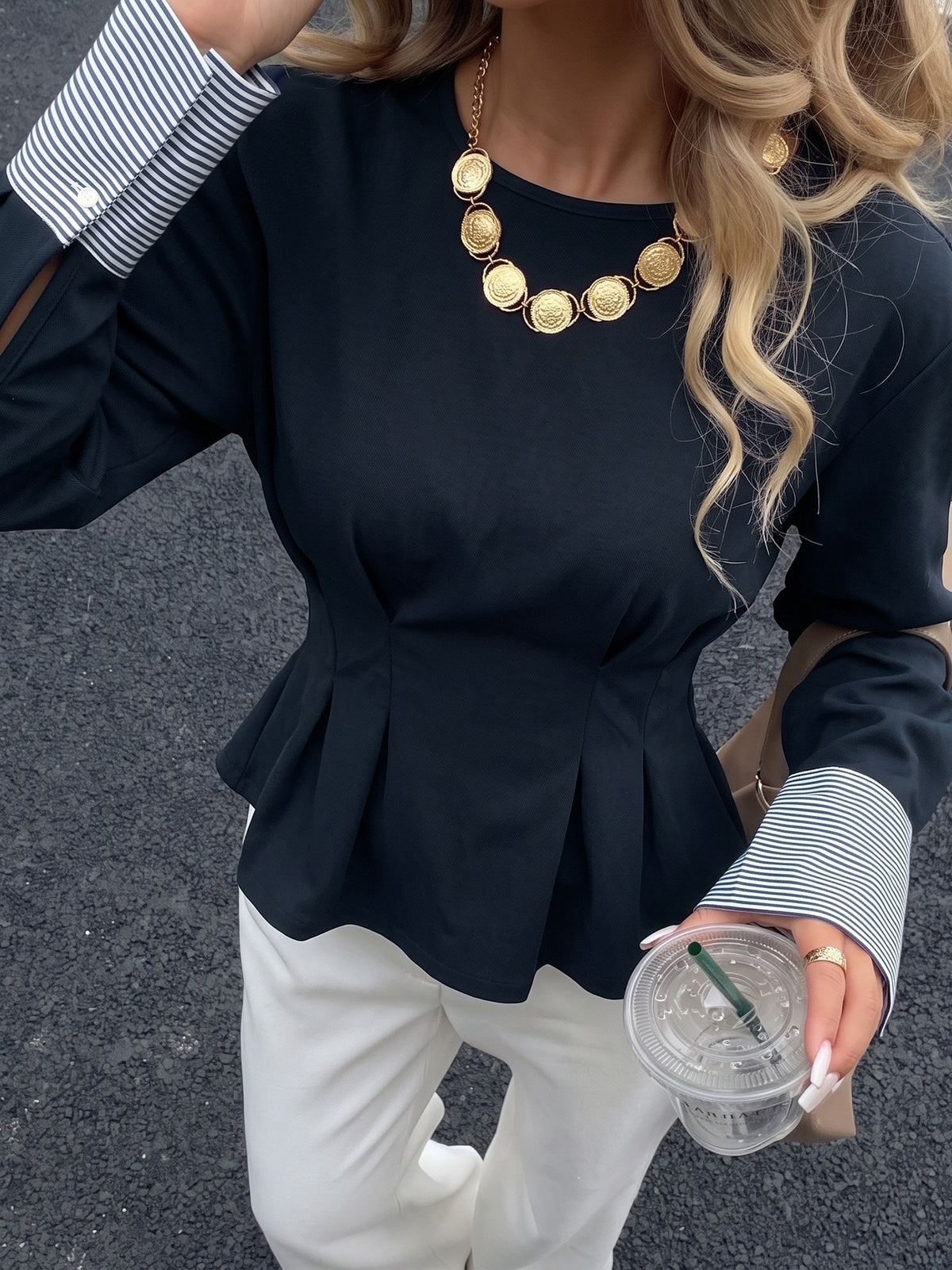 A woman wearing a black long-sleeve peplum top with striped buttoned cuffs, paired with white pants and a gold coin necklace, holding a clear plastic cup on a dark asphalt surface.