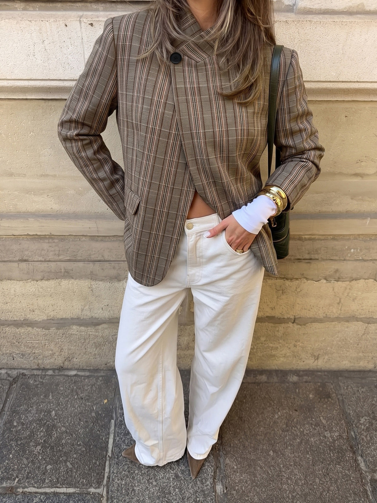 A woman wearing a brown and beige plaid wrap-style blazer, white wide-leg trousers, and brown pointed-toe heels, standing against a beige stone wall.