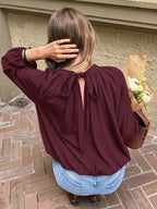 A woman viewed from behind wearing a long-sleeve burgundy blouse with a tie-back neckline, light blue jeans, and leopard-print shoes, while holding a brown paper bag with white flowers against a beige stucco wall and brick patio.