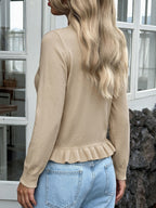 A woman shown from the back wearing a beige long-sleeve knit top with a ruffled hem, paired with light blue jeans, standing in front of a white-framed window.