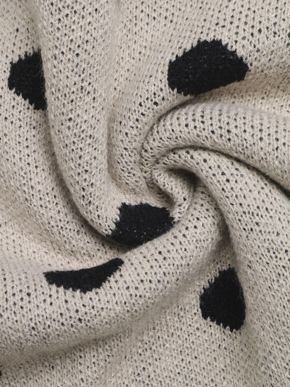 Close-up of a knitted fabric in a warm off-white or cream tone with irregular black spots, resembling a dalmatian-style pattern.