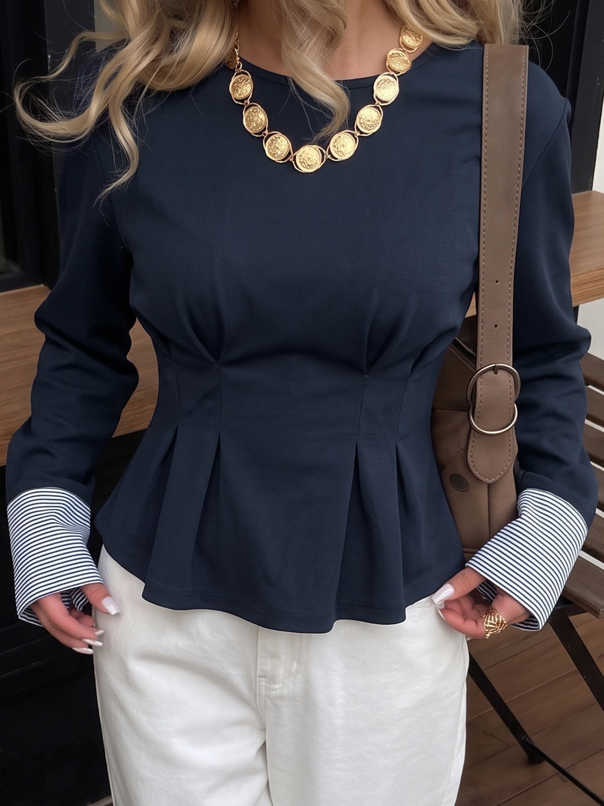 A woman wearing a navy blue long-sleeve peplum top with striped buttoned cuffs, paired with white pants and a chunky gold coin necklace. She is carrying a brown shoulder bag.
