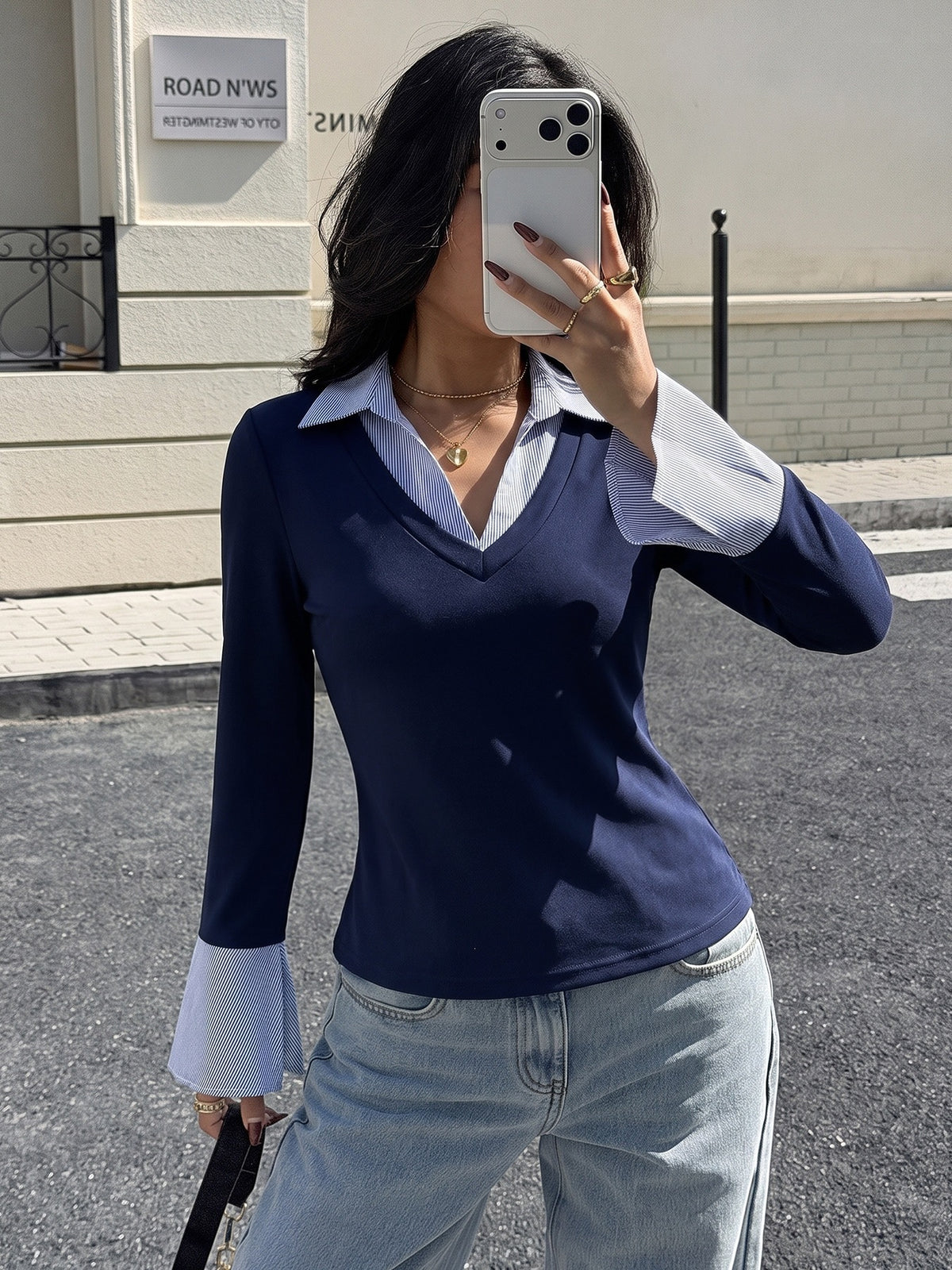 A woman takes a mirror selfie wearing a navy blue long-sleeve V-neck top with light blue bell-sleeve undersleeves, paired with light-wash jeans.