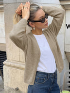 A woman wearing a beige ribbed knit cardigan over a white t-shirt and blue jeans, posing outdoors against a stone wall.