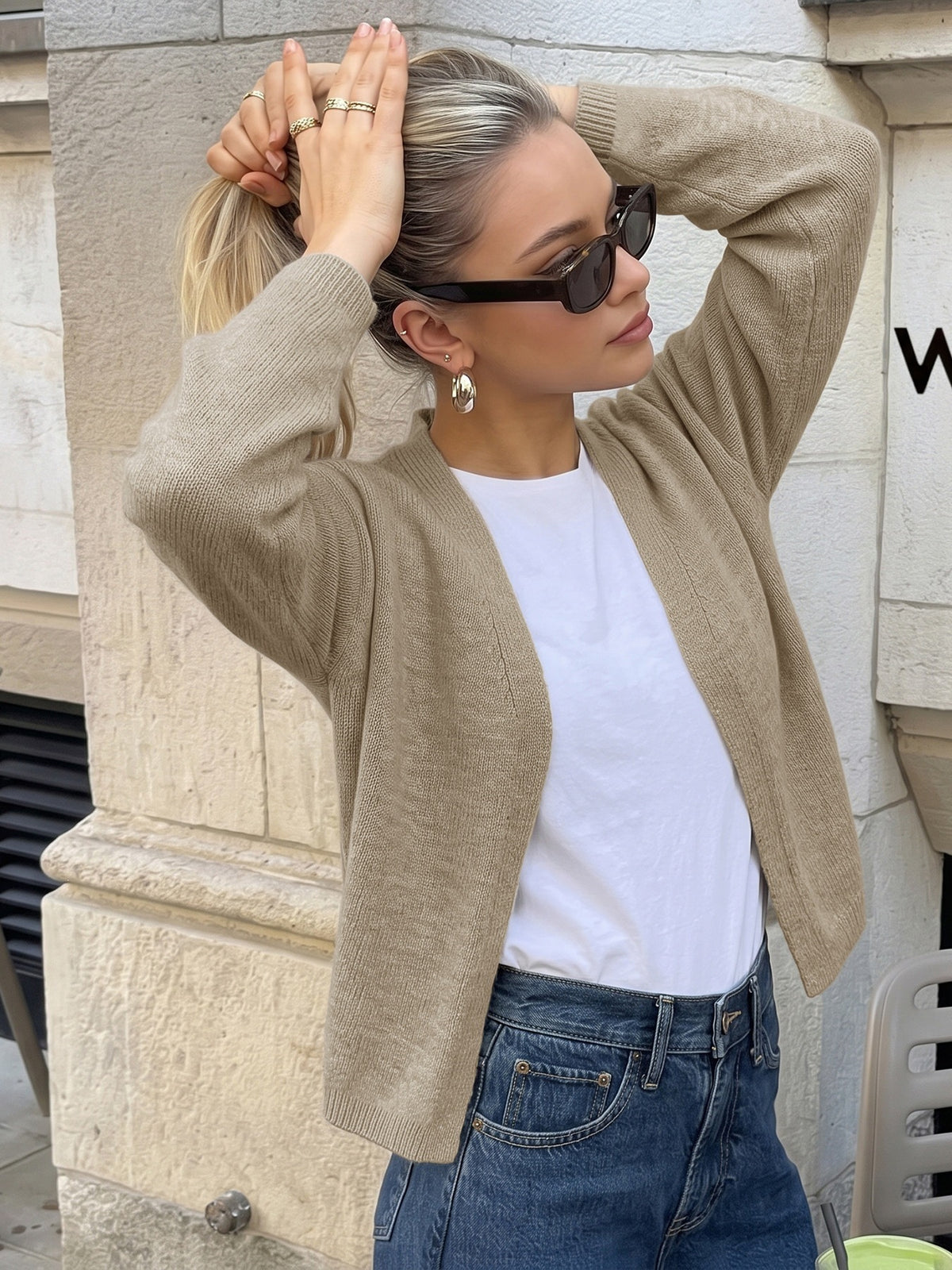 A woman wearing a beige ribbed knit cardigan over a white t-shirt and blue jeans, posing outdoors against a stone wall.