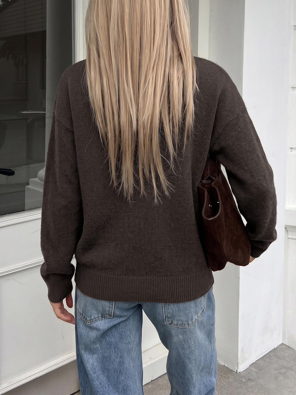 A person viewed from behind wearing a dark brown knit sweater and light blue jeans, holding a brown shoulder bag against a white building exterior.