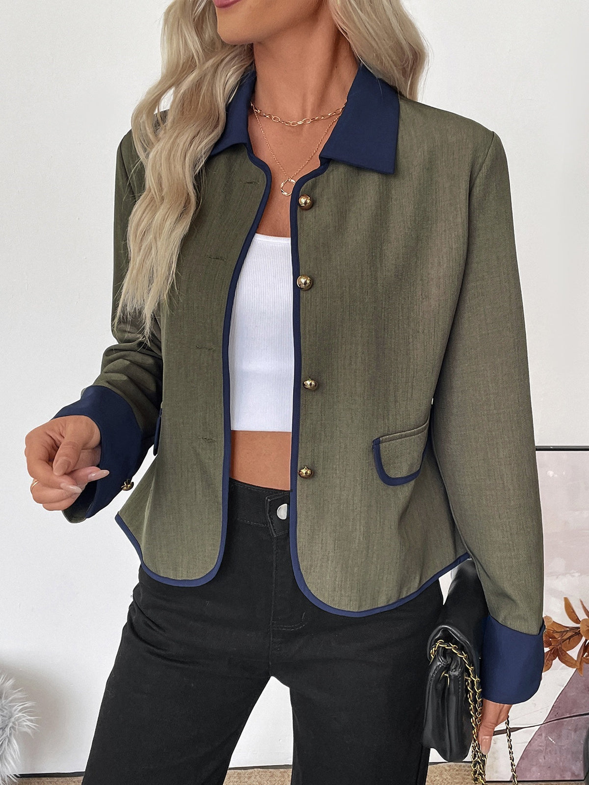 A woman wearing a cropped olive green blazer with contrasting navy blue collar, cuffs, and hem trim, styled with a white crop top and black pants.
