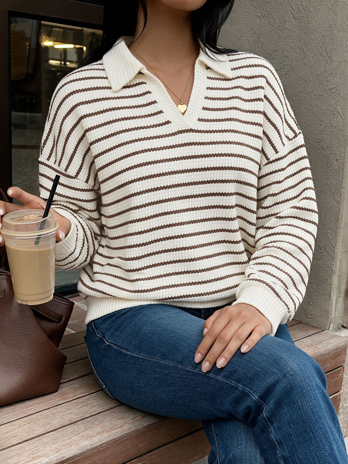 A woman wearing a cream and brown horizontally striped knit polo sweater sits on a wooden bench, holding an iced coffee.