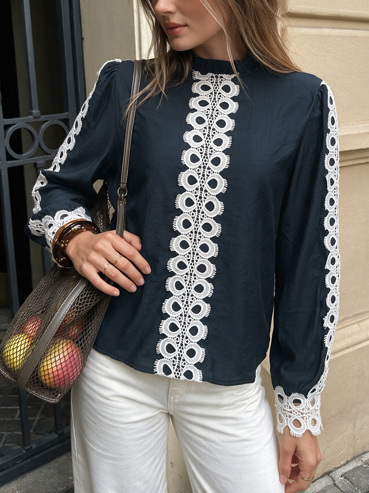 A woman wearing a navy blue long-sleeve blouse with prominent white lace trim down the front and along the sleeves, paired with white pants and carrying a brown mesh shoulder bag.