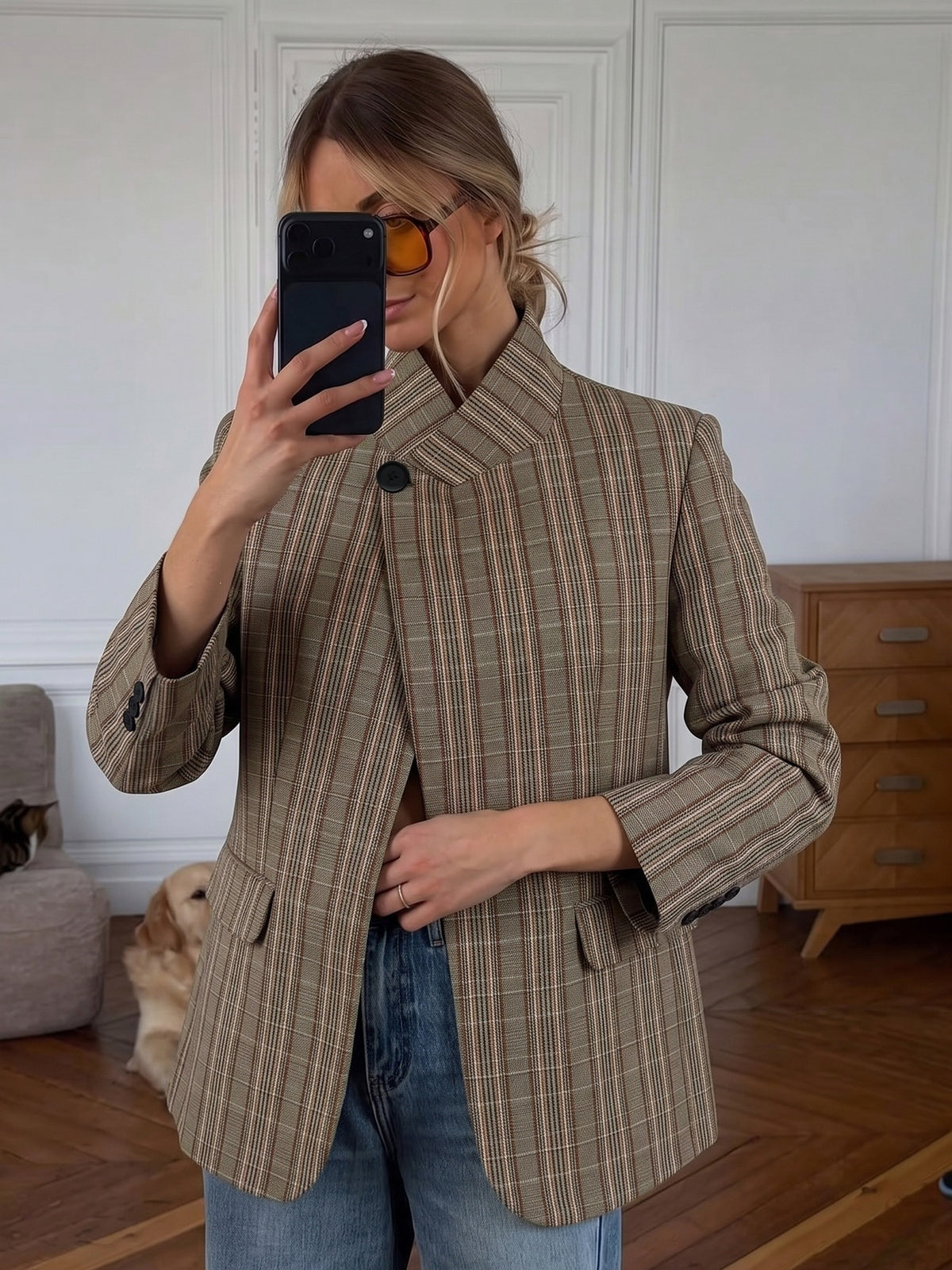 A woman takes a mirror selfie wearing a brown and beige plaid blazer with blue jeans in a home setting with a wooden floor and a wooden chest in the background.