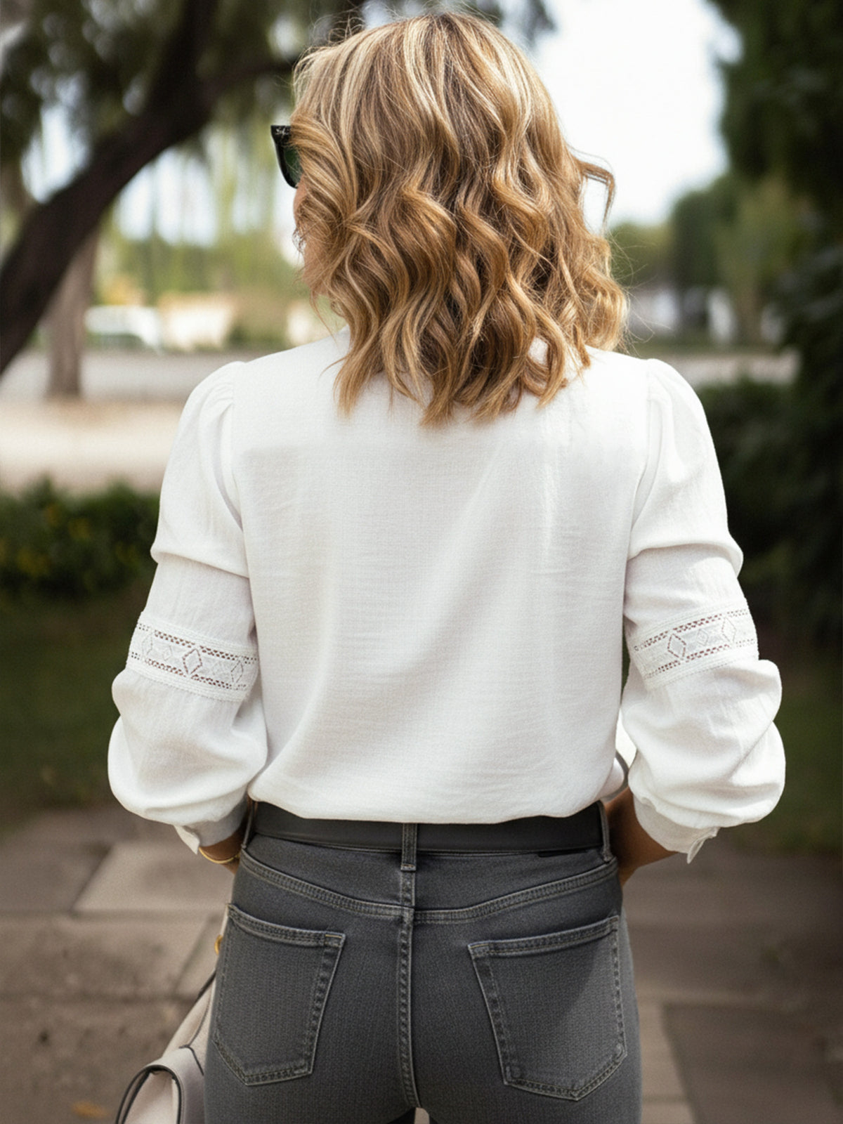 A woman viewed from behind wearing a white long-sleeve blouse with lace sleeve detailing and dark gray jeans, standing outdoors on a paved path with trees in the background.