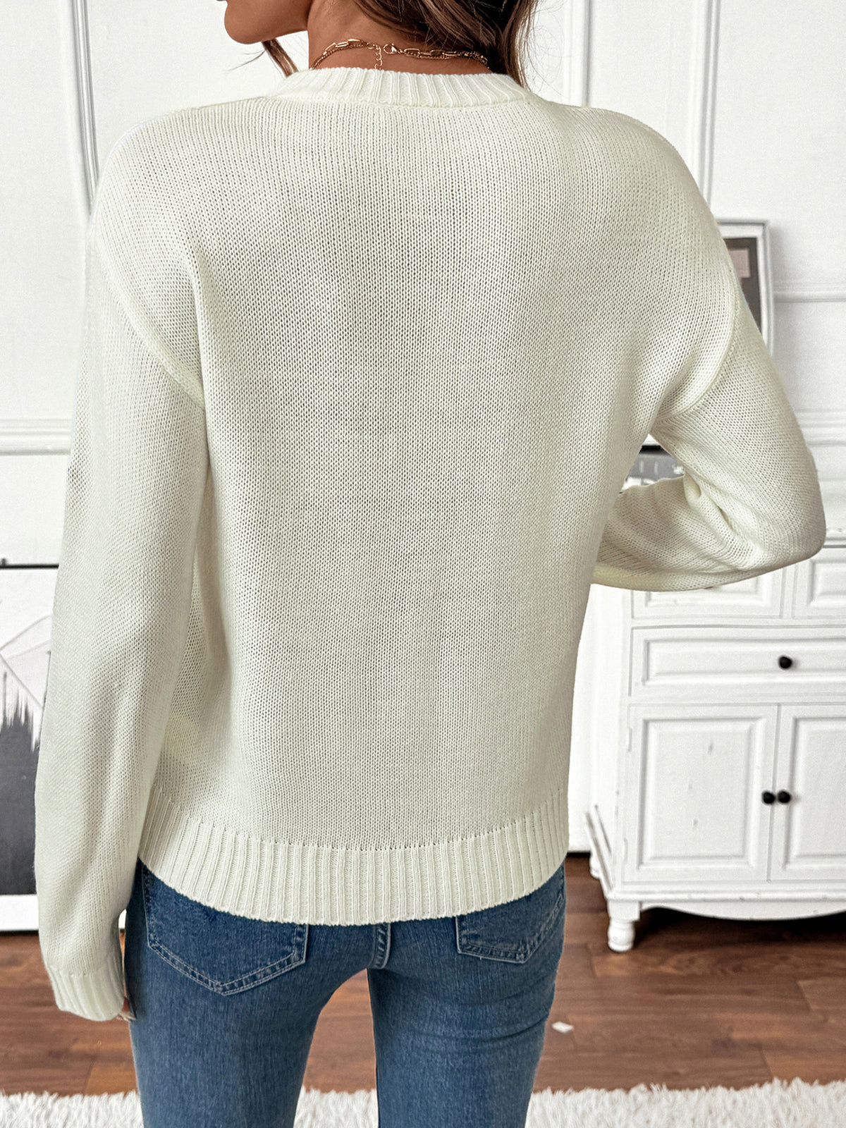 A woman shown from the back wearing a cream-colored knit sweater and blue jeans in a bright indoor room with white furniture.