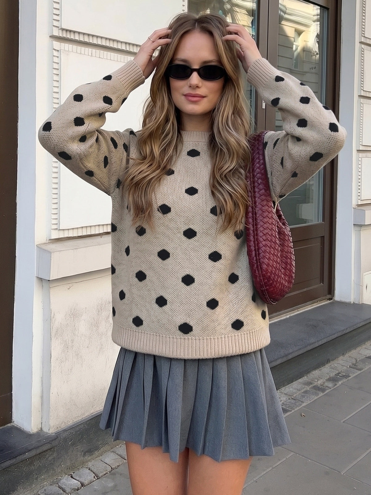 A woman wearing a beige knit sweater with black polka dots and a gray pleated skirt, standing outdoors in front of a building with a brown door. She is wearing black sunglasses and carrying a burgundy woven shoulder bag.
