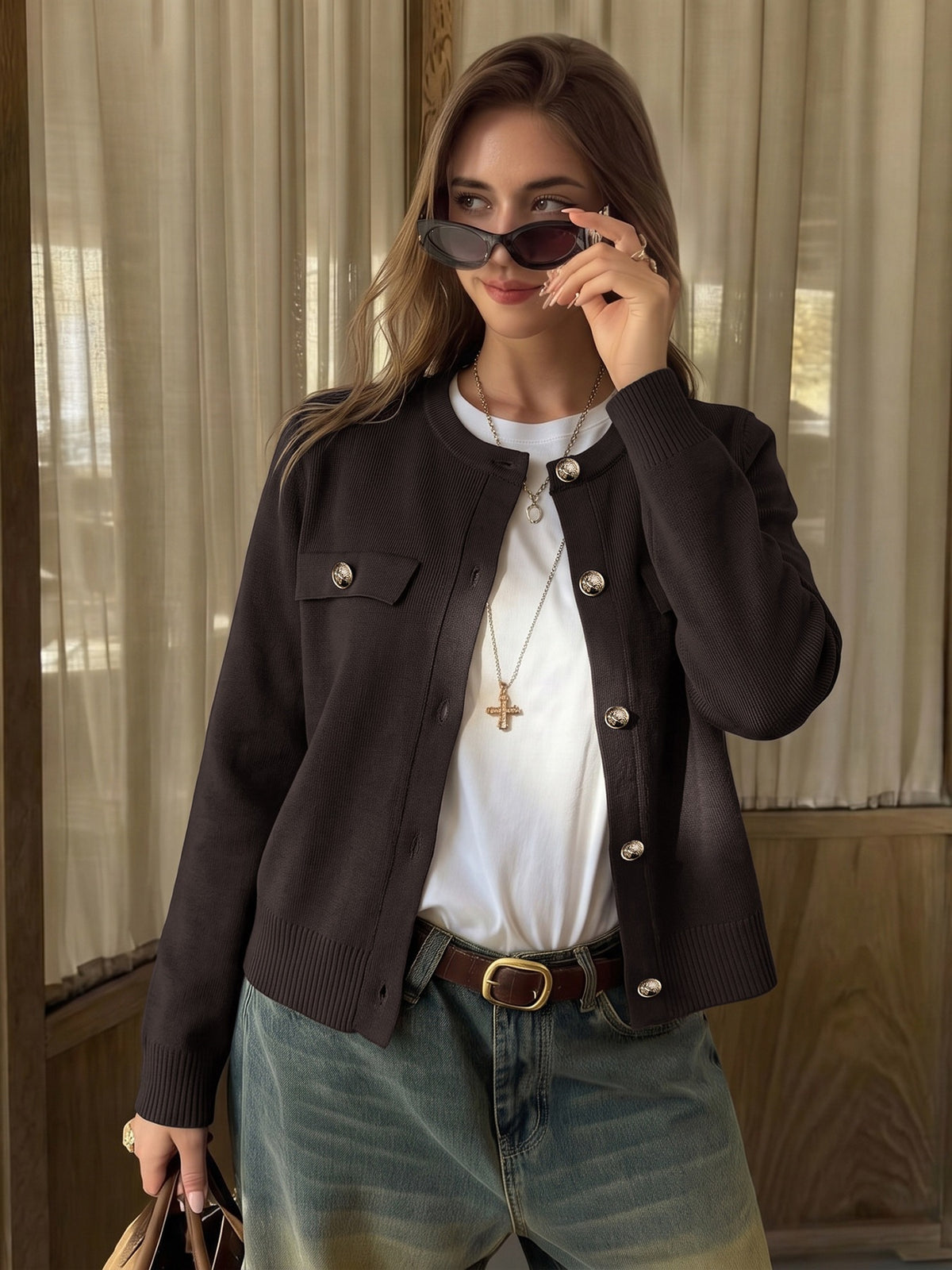 A woman wearing a dark brown ribbed knit cardigan with silver buttons over a white t-shirt, paired with blue jeans and holding a brown handbag, set against a background of beige curtains.