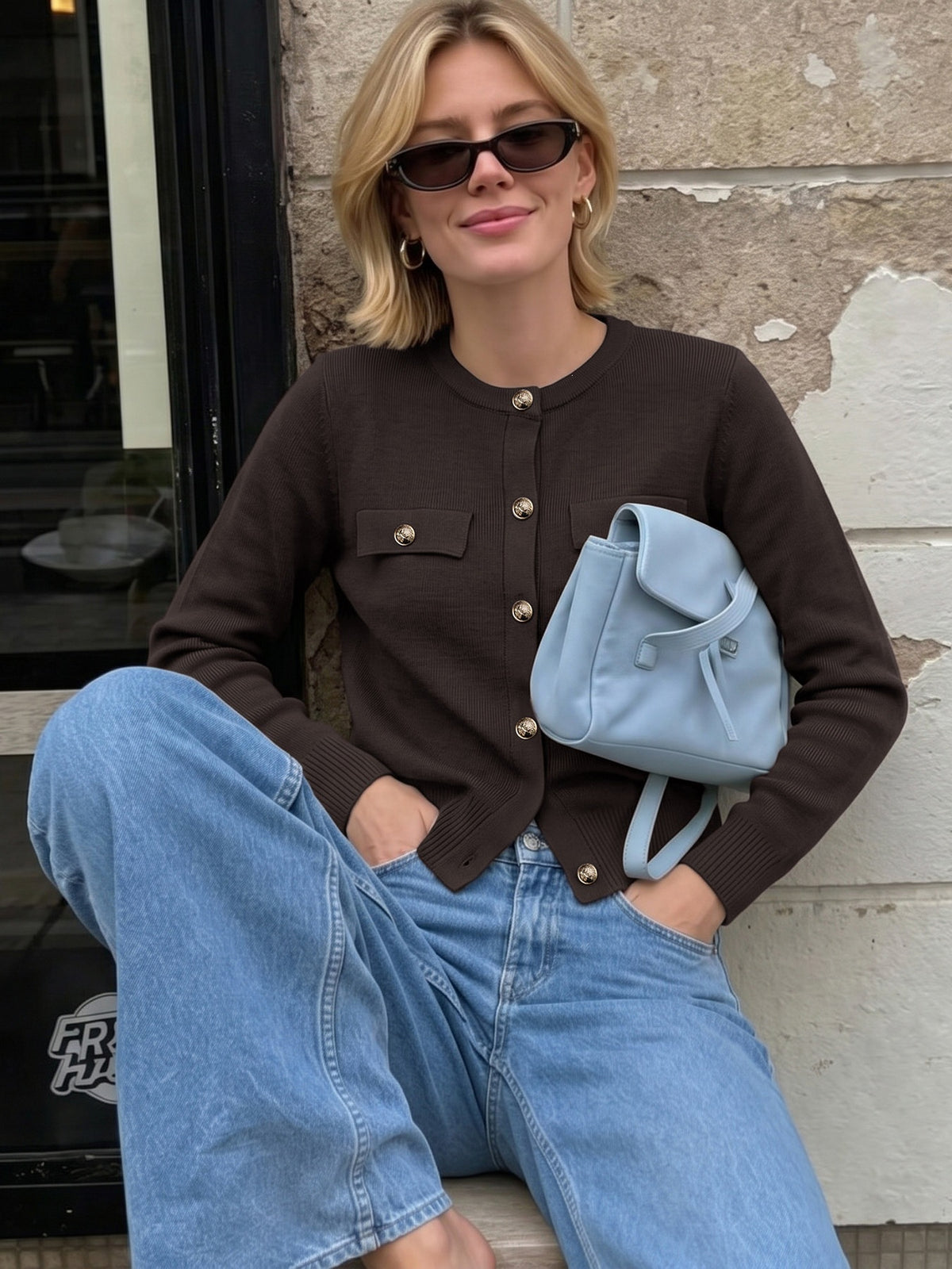 A woman wearing a dark brown button-up cardigan, light blue jeans, and sunglasses, holding a light blue leather bag against a stone wall background.