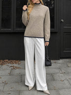 A woman wearing a beige knit mock-neck sweater with dark trim, white wide-leg trousers, and beige pointed-toe heels, holding a small black handbag while standing outdoors in front of a dark building.