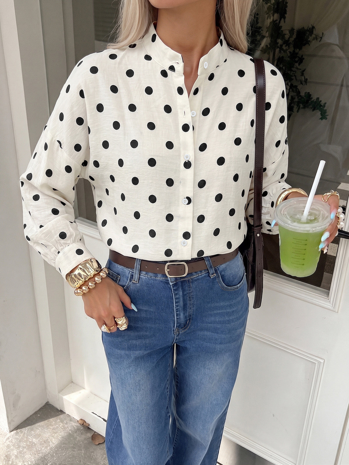 A woman wearing a white long-sleeve button-up blouse with black polka dots, paired with blue jeans and a brown belt, holding a green drink.