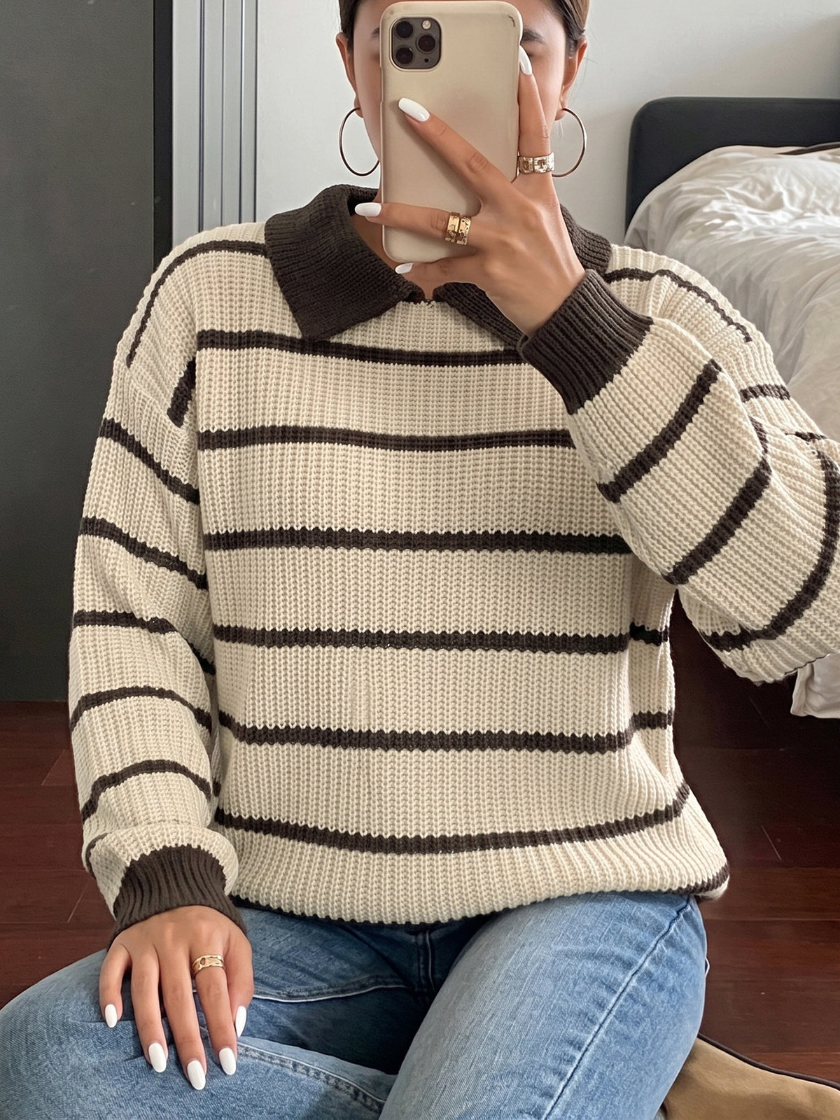 A person taking a mirror selfie wearing a cream and dark brown horizontally striped knit sweater with a dark brown collar and cuffs, paired with blue jeans.