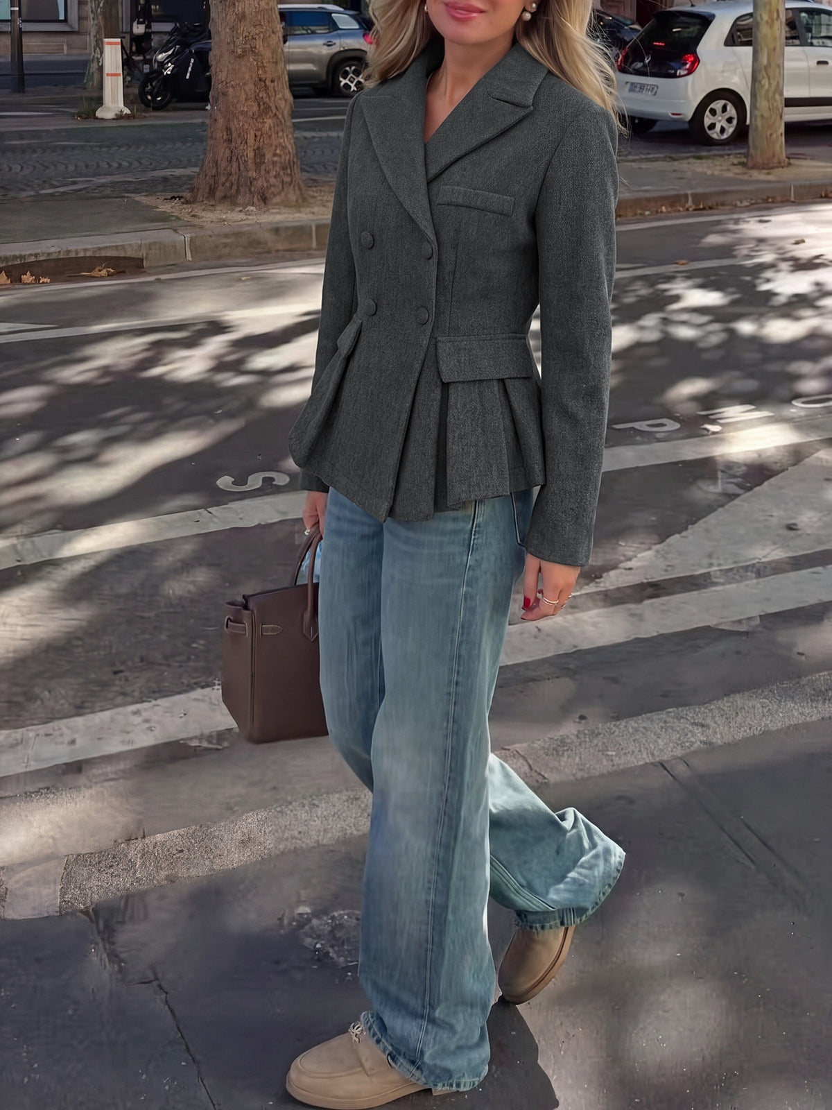 A woman wearing a dark gray double-breasted peplum blazer, light-wash wide-leg jeans, and beige sneakers, carrying a brown handbag while walking on a city street crosswalk.