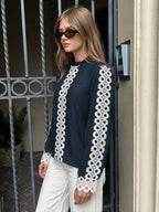 A woman wearing a black long-sleeve blouse with intricate white lace trim down the front and along the sleeves, paired with white pants and black sunglasses, standing in front of a metal gate.