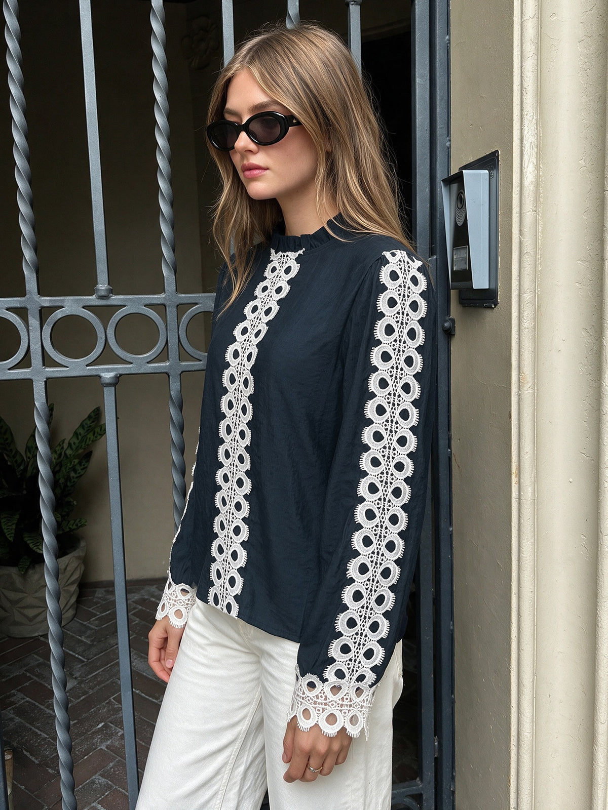A woman wearing a black long-sleeve blouse with intricate white lace trim down the front and along the sleeves, paired with white pants and black sunglasses, standing in front of a metal gate.
