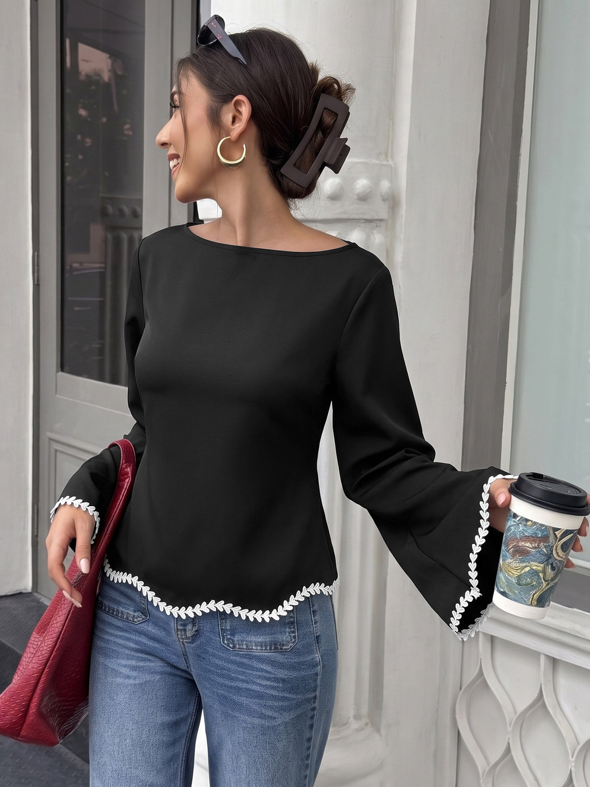 A woman wearing a black long-sleeve top with white decorative trim along the hem and cuffs, paired with blue jeans, holding a red handbag and a coffee cup in front of a white building.