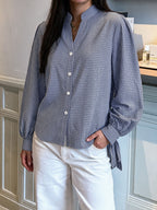 A woman wearing a blue and white gingham long-sleeve button-up shirt with a stand collar and side-tie detail, paired with white pants, standing indoors.