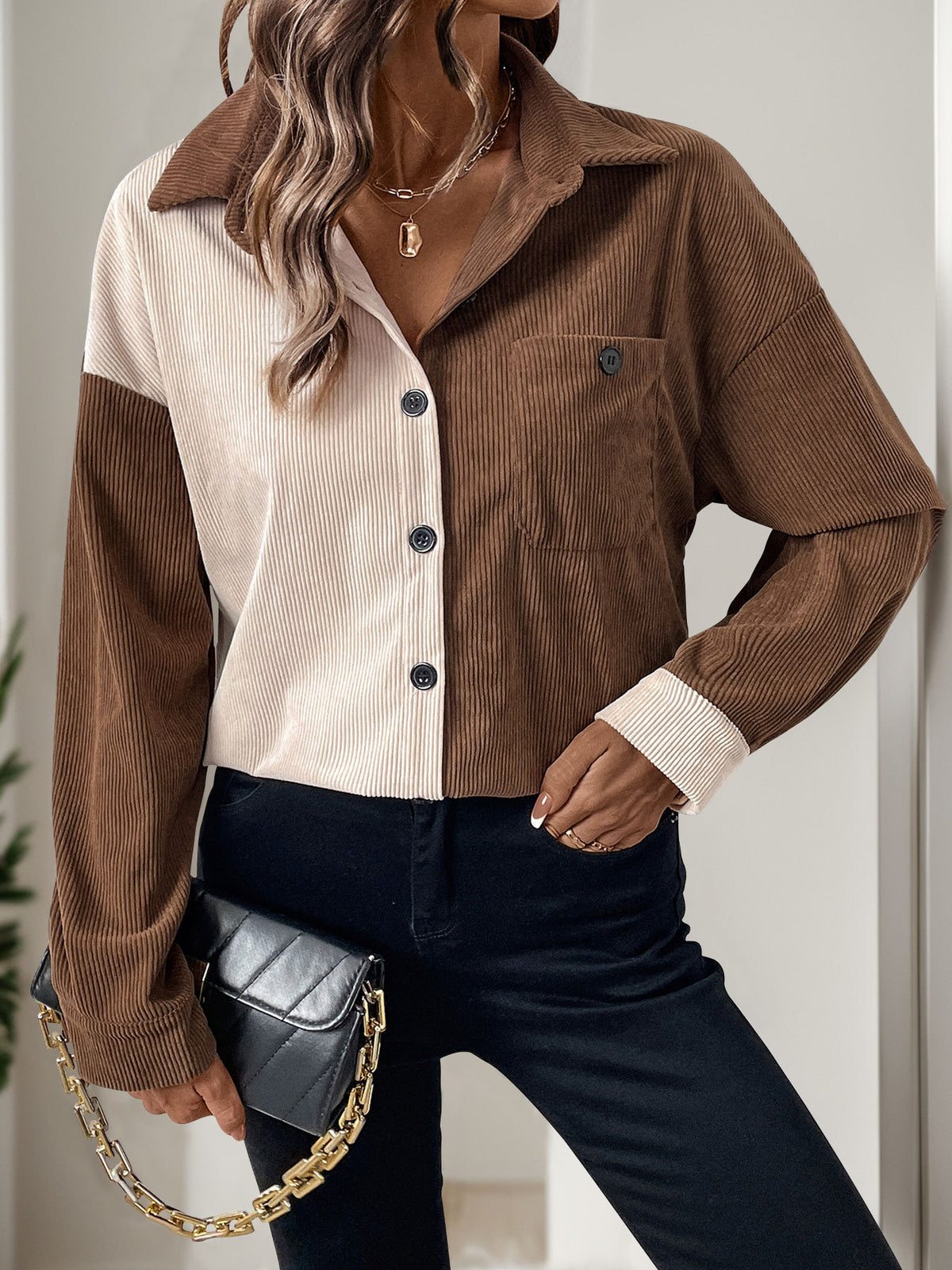 A woman models a color-block corduroy button-up shirt with brown sleeves and collar, and a cream ribbed body. She is wearing dark blue jeans and holding a black quilted handbag with a gold chain strap.