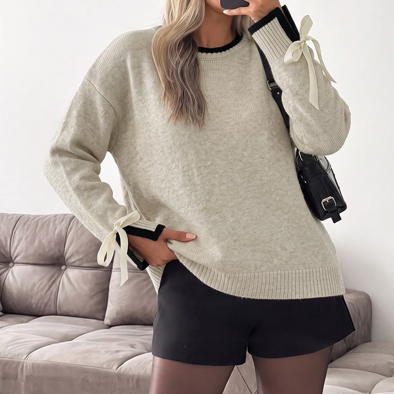 A woman wearing an oversized light beige knit sweater with black trim and cream ribbon ties at the cuffs, paired with black shorts and a black shoulder bag, standing in front of a gray tufted sofa.