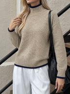 A woman wearing a beige knit mock neck sweater with navy blue ribbed trim at the collar, cuffs, and hem, paired with white trousers and a black shoulder bag.