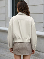 A woman viewed from behind wearing a light beige cropped jacket and a brown mini skirt, standing outdoors against a light gray building wall.