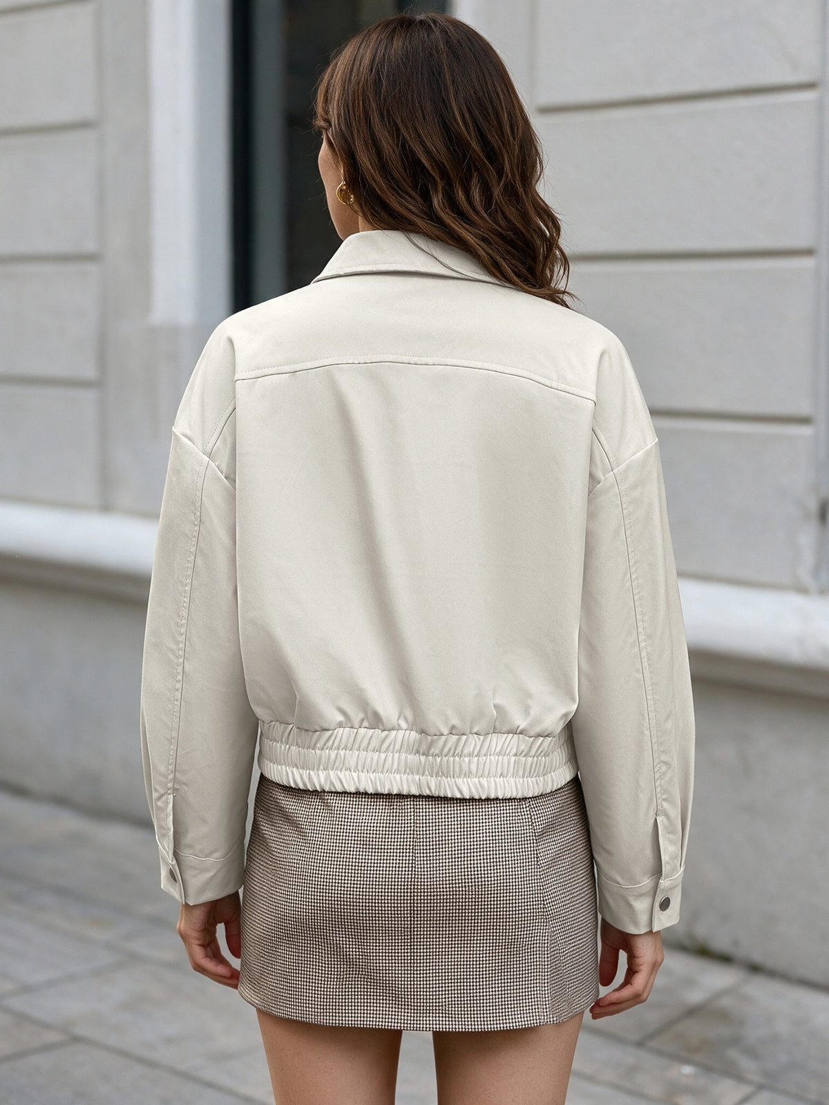 A woman viewed from behind wearing a light beige cropped jacket and a brown mini skirt, standing outdoors against a light gray building wall.