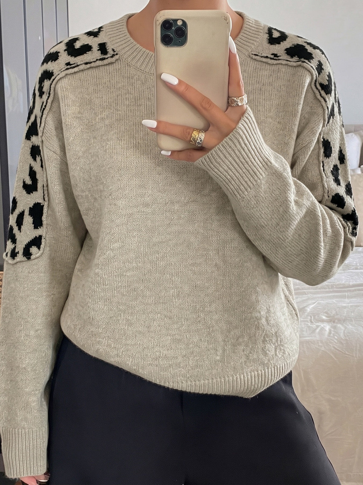 A person takes a mirror selfie wearing a beige knit sweater with black leopard print shoulder panels, paired with black pants.