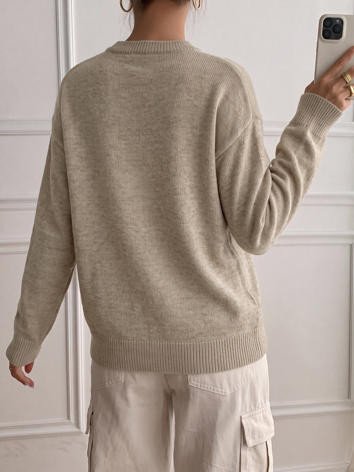 A person shown from the back wearing a light beige knit sweater and matching light cargo pants, standing indoors against a white paneled wall while holding a phone.